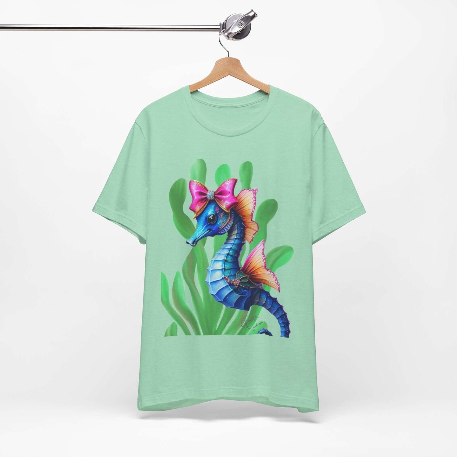 Mint green T-shirt on a wooden hanger featuring a colorful seahorse illustration with a large pink bow and pastel fins, set against soft green seaweed. Perfect for marine-themed fashion, cute sea creature lovers, and bold aquatic apparel collections.