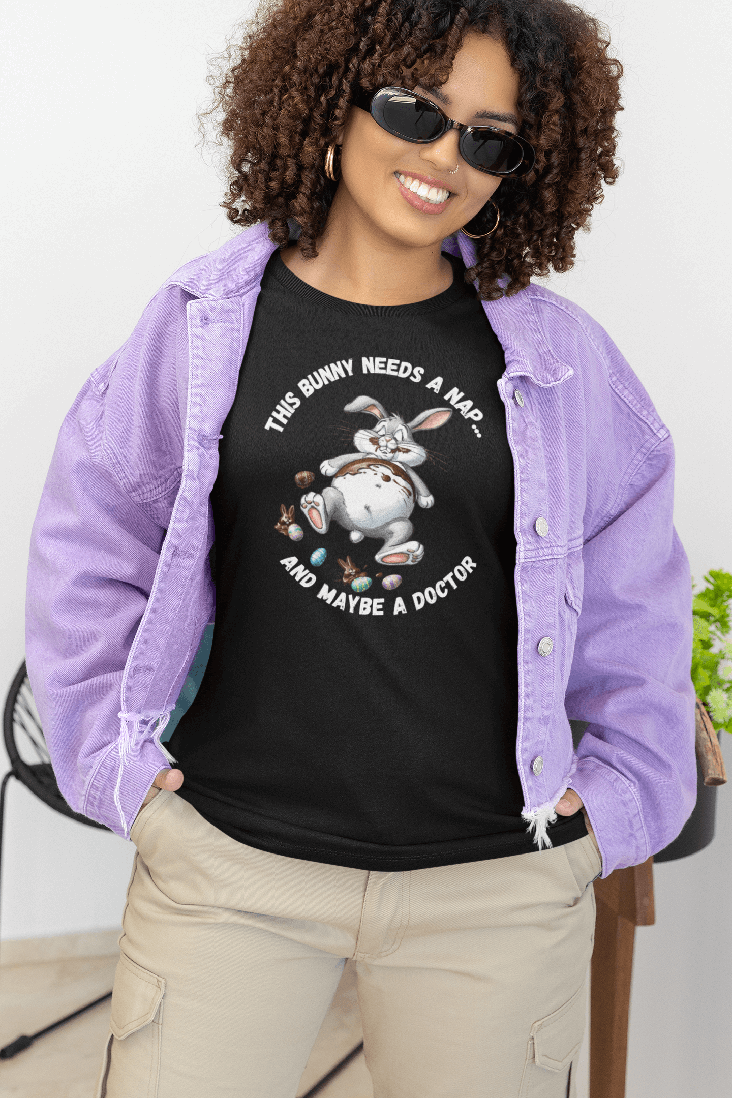 Woman wearing black tee with cartoon bunny lying flat and surrounded by chocolate, with the text “This Bunny Needs a Nap... And Maybe a Doctor.” Styled with lilac jacket—perfect for Easter humor and funny spring T-shirt lovers.