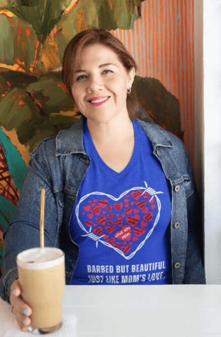 Smiling woman in a royal blue v-neck t-shirt with a white barbed wire heart filled with red hearts labeled with affectionate mom phrases, plus the quote “Barbed but beautiful, just like Mom’s love.” Bold apparel for coffee-loving queens of quirky fashion.