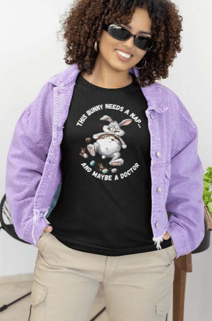Woman wearing a black t-shirt with a graphic of an exhausted cartoon bunny lying on its back, surrounded by chocolate and candy. The caption reads “This Bunny Needs a Nap... and Maybe a Doctor.” Styled with a lilac jacket and beige cargo pants, this look blends humor, comfort, and spring fashion. Perfect for Easter, chocolate lovers, or introverts on the edge.
