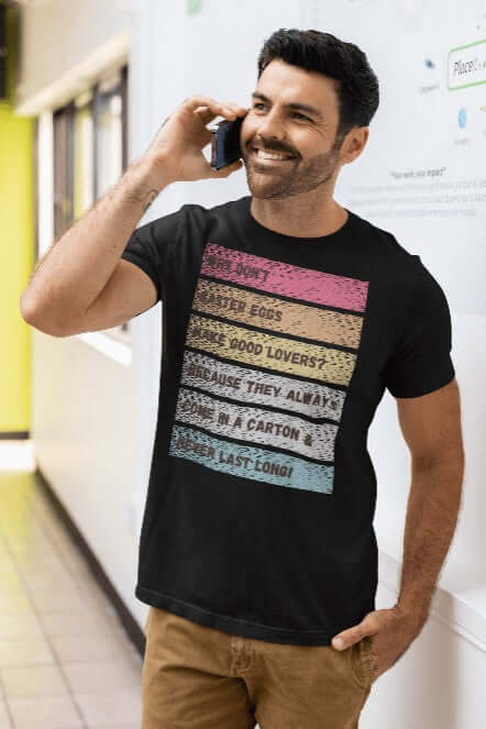 Smiling man in a black t-shirt standing in a hallway, talking on the phone. His tee features a distressed retro rainbow design with a bold white caption: “Why don’t Easter eggs make good lovers? Because they always come in a carton & never last long!” A hilarious Easter graphic tee perfect for adults, party hosts, and pun enthusiasts.