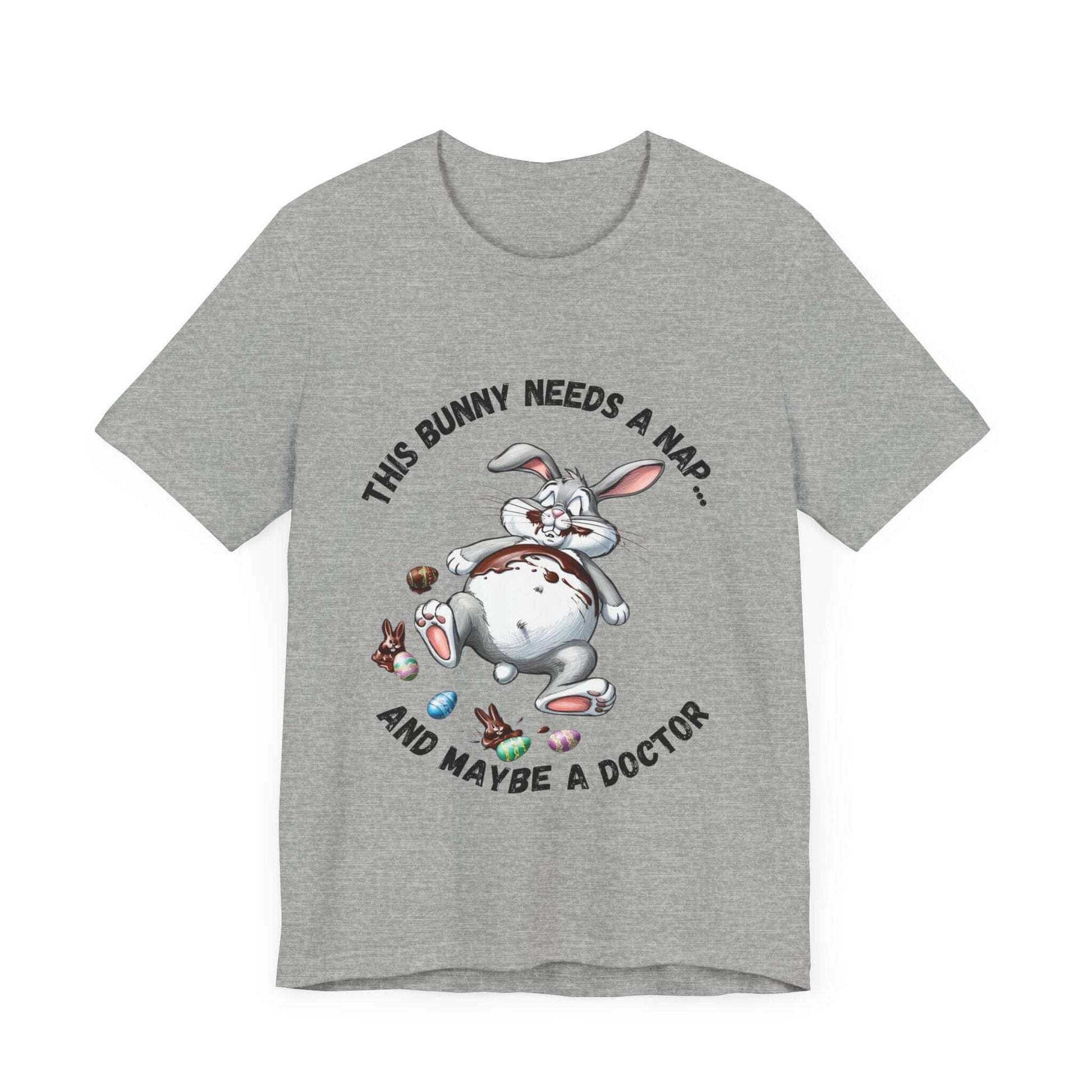 Light grey t-shirt featuring a chubby cartoon bunny sprawled out belly-up, surrounded by candy wrappers and melty chocolate. The text reads “This Bunny Needs a Nap... and Maybe a Doctor.” A perfect Easter humor tee for tired snackers and chocolate lovers.