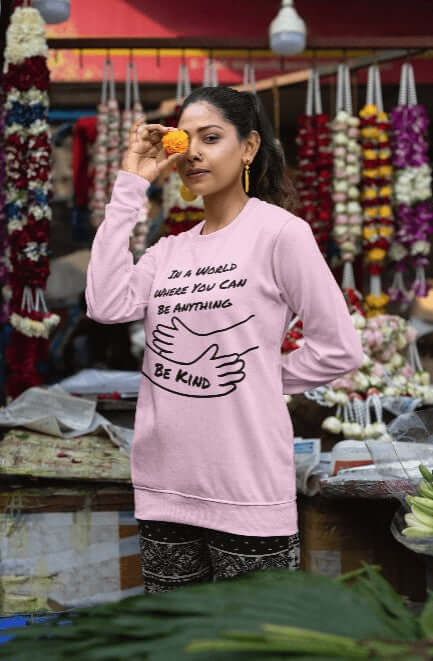 Woman standing in a vibrant flower market, wearing a light pink sweatshirt featuring the phrase “In a world where you can be anything, be kind” with a black line art graphic of two hugging arms. The design promotes kindness, emotional wellness, and mental health awareness in a cozy and stylish crewneck — ideal for everyday wear, teachers, therapists, and self-care advocates.