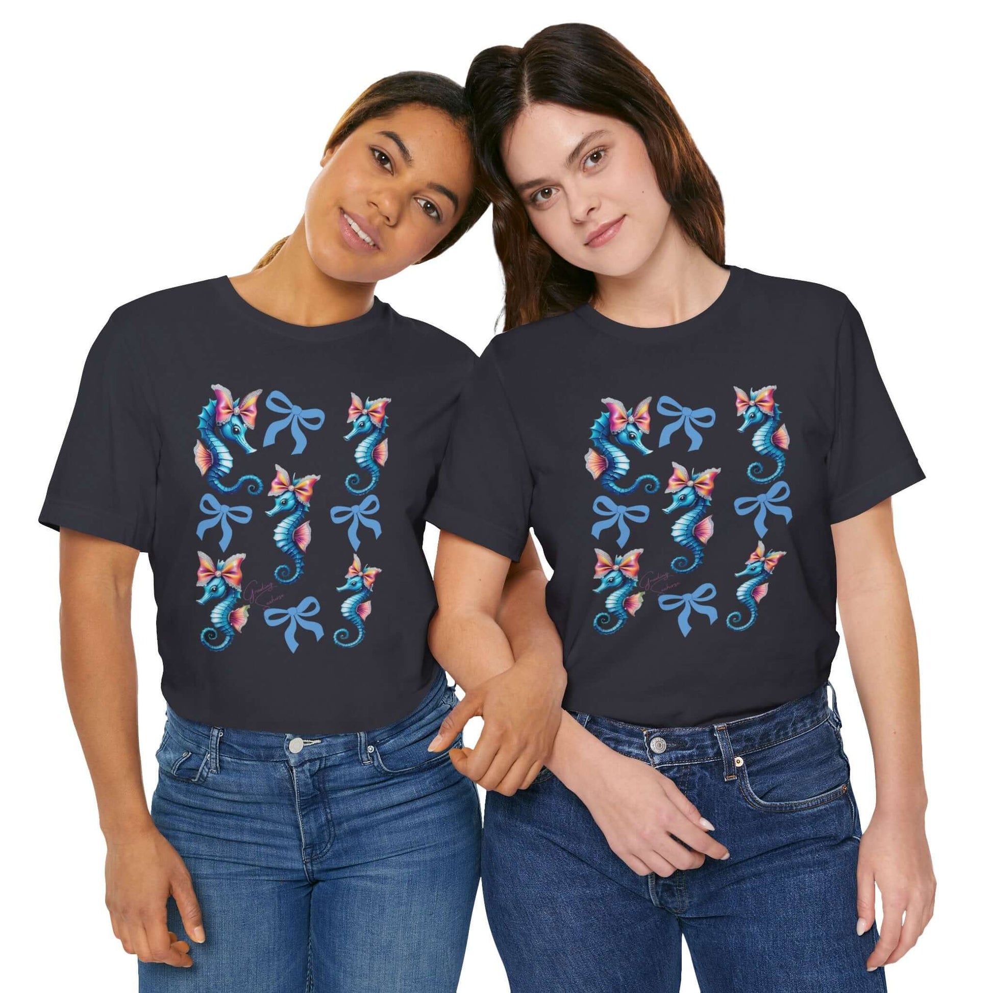 Two smiling women wearing matching dark grey t-shirts featuring a pastel seahorse and bow graphic arranged in a symmetrical grid. The design includes soft pink bows and vintage-style sea creatures, blending cottagecore, coquette fashion, and ocean whimsy into a bold apparel look.