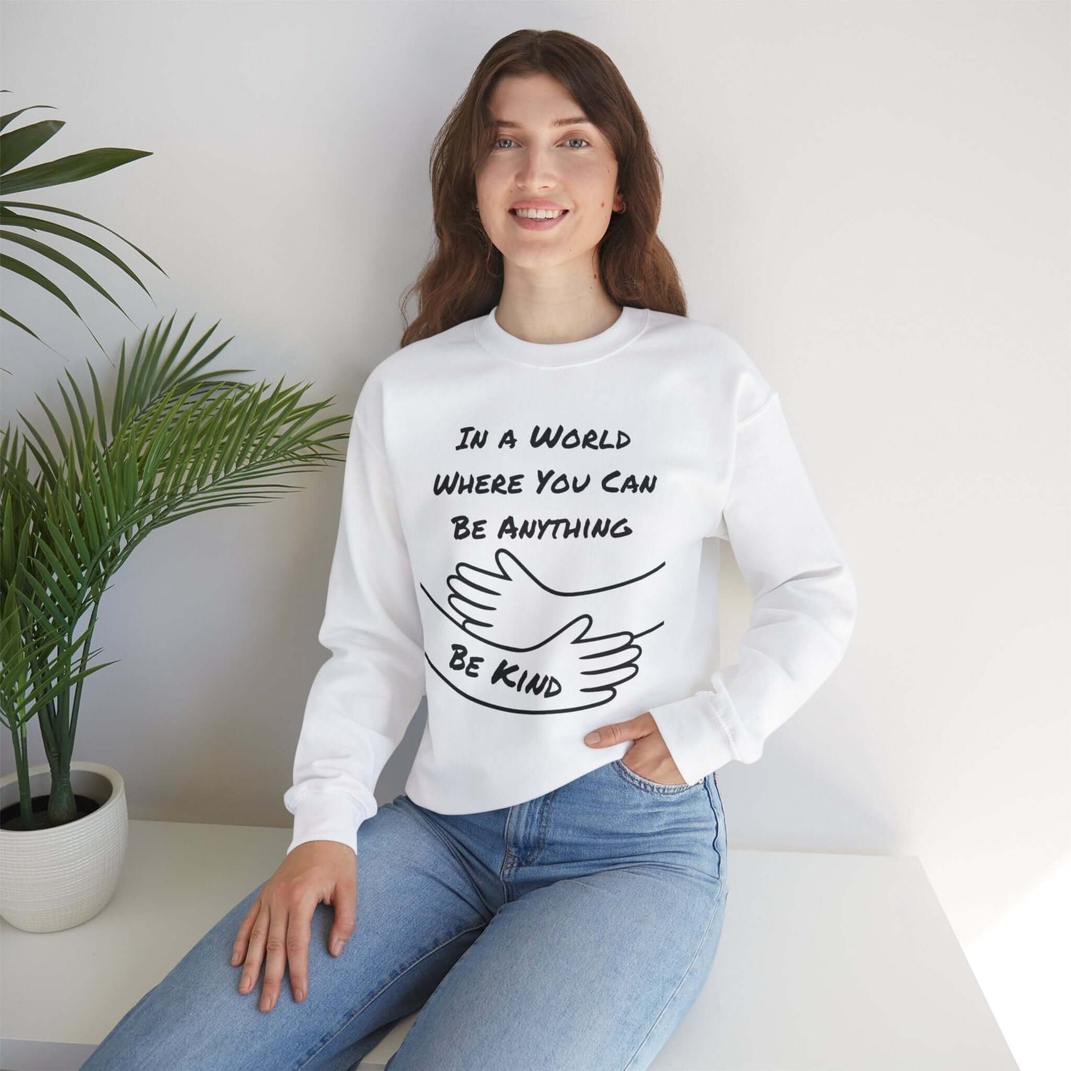 Woman wearing white sweatshirt with hugging arms graphic and the message “In a world where you can be anything, be kind.” Casual, cozy style perfect for self-care, mental health awareness, and kindness-themed apparel collections.
