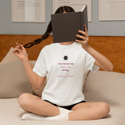 Person sitting on a couch with a book covering their face, wearing a white t-shirt with text.