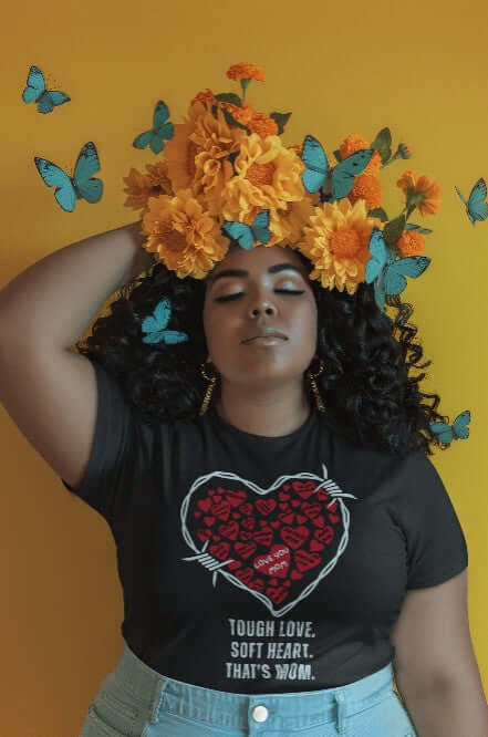 Confident woman wearing a black t-shirt with a white barbed wire heart filled with red hearts and loving mom phrases. Surrounded by yellow flowers and blue butterflies. Bold apparel meets quirky fashion in this artistic Mother's Day tribute tee.