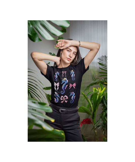 Woman standing among tropical plants, wearing a black t-shirt with a colorful design of nine pastel seahorses and four oversized bows. The layout is symmetrical and playful, evoking a dreamy, whimsical undersea world. Perfect for fans of ocean-themed apparel, soft girl fashion, and Coquette-core vibes.