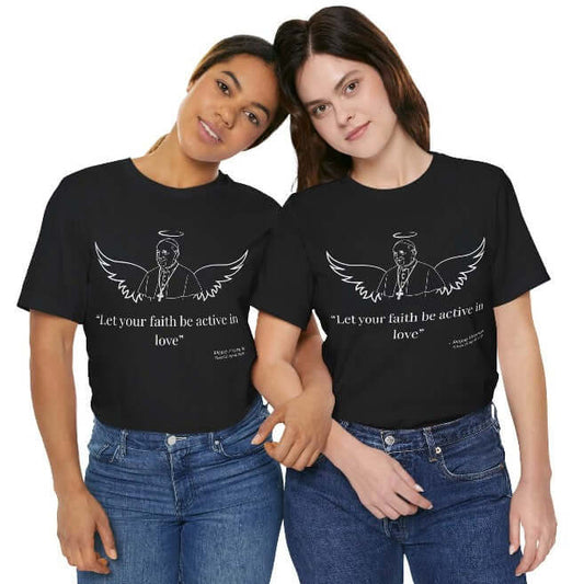 Two women wearing black unisex Pope Francis tribute T-shirts featuring angel wings, halo, minimalist line art portrait, and quote: “Let your faith be active in love.”