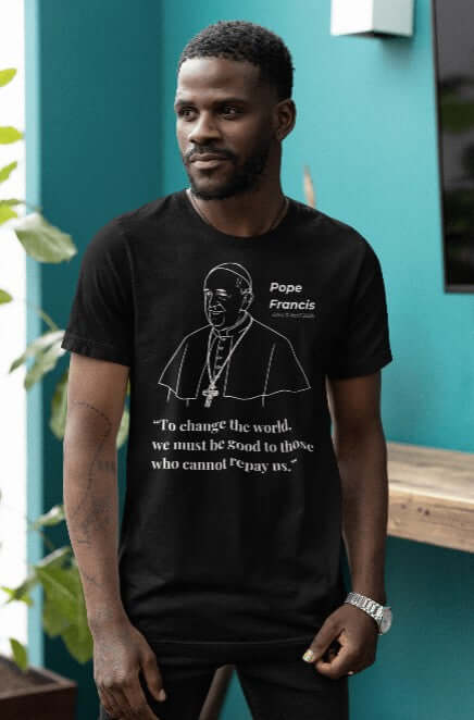 Man wearing a black t-shirt featuring a minimalist white line art portrait of Pope Francis with a halo and cross necklace. Below the image, the quote reads: “To change the world, we must be good to those who cannot repay us.” A Catholic memorial shirt celebrating compassion, humility, and the legacy of The People’s Pope.