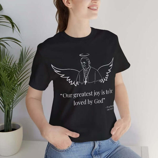 Smiling woman wearing a black t-shirt with a white minimalist line art of Pope Francis featuring angel wings and a halo. Text below reads: “Our greatest joy is to be loved by God.” A heartfelt Catholic tribute shirt honoring the legacy of Pope Francis with grace and spiritual peace.