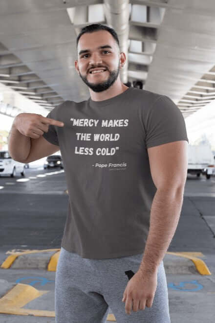 Man smiling and pointing at his dark grey t-shirt featuring the quote “Mercy makes the world less cold” – Pope Francis (Died 2 April 2025). The design uses minimalist white typography and is styled as a Catholic memorial shirt that honors compassion, faith, and servant leadership.