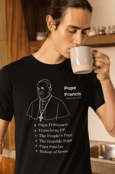 Man drinking from a mug while wearing a black t-shirt featuring a white line art portrait of Pope Francis, halo detail, and a list of names including “Papa Francesco,” “Franciscus PP.,” “The People’s Pope,” and “Bishop of Rome.” A respectful Catholic memorial shirt paying tribute to his faith, leadership, and humble legacy.