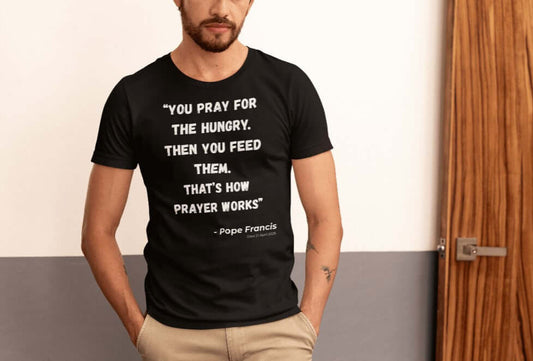Man wearing a black t-shirt with bold white text reading “You pray for the hungry. Then you feed them. That’s how prayer works” – Pope Francis (Died 2 April 2025). A minimalist Catholic tribute tee that honors Pope Francis’s message of service, compassion, and action through faith.