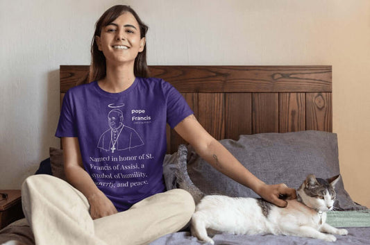 Smiling woman sitting on a bed, wearing a purple t-shirt with a white minimalist line art of Pope Francis with a halo. Text below reads: “Named in honor of St. Francis of Assisi, a symbol of humility, poverty, and peace.” A spiritual, commemorative tee perfect for Catholic memorial or reflection.