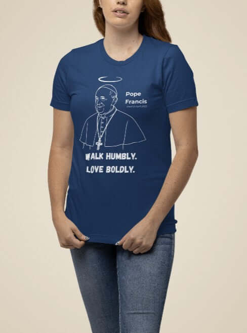 Woman wearing a navy blue t-shirt featuring a white minimalist line art portrait of Pope Francis with a halo and cross necklace, alongside bold text that reads “Walk Humbly. Love Boldly.” A Catholic memorial shirt honoring his compassionate, faith-filled leadership.