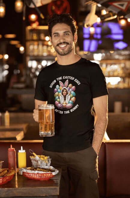 Man holding a beer and smiling in a restaurant setting while wearing a black t-shirt with a cheeky Easter graphic. The design shows a woman in a bunny costume surrounded by Easter eggs, paired with the caption “Screw the Easter Eggs, I’m After the Bunny.” A flirty, funny shirt for bold holiday gatherings or adults with a sense of humor.
