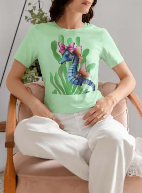 Woman seated in a soft pink chair wearing a mint green t-shirt featuring a pastel blue seahorse with coral-colored fins and a large sparkly pink bow, nestled in leafy green seaweed. A whimsical, coquette-style design perfect for fans of soft girl fashion, oceancore, and bold apparel with aesthetic charm.