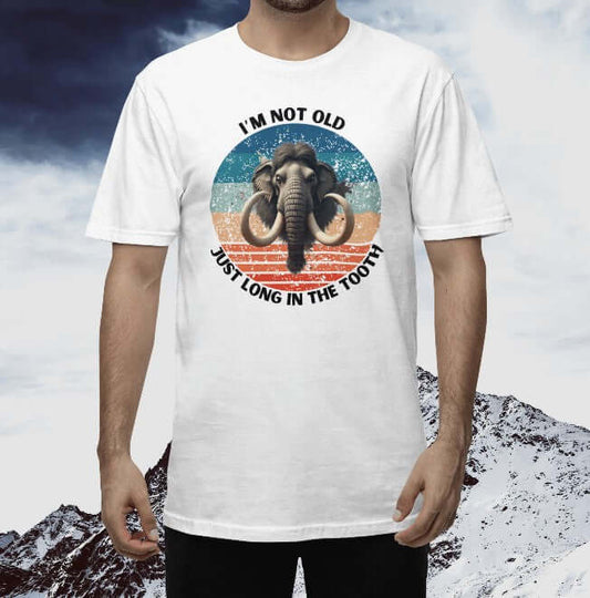 Man wearing a white unisex T-shirt featuring a bold graphic of a woolly mammoth centered in a retro sunset circle with the phrase “I’m not old, just long in the tooth.” A funny age-themed gift tee with vintage tusks and prehistoric vibes.